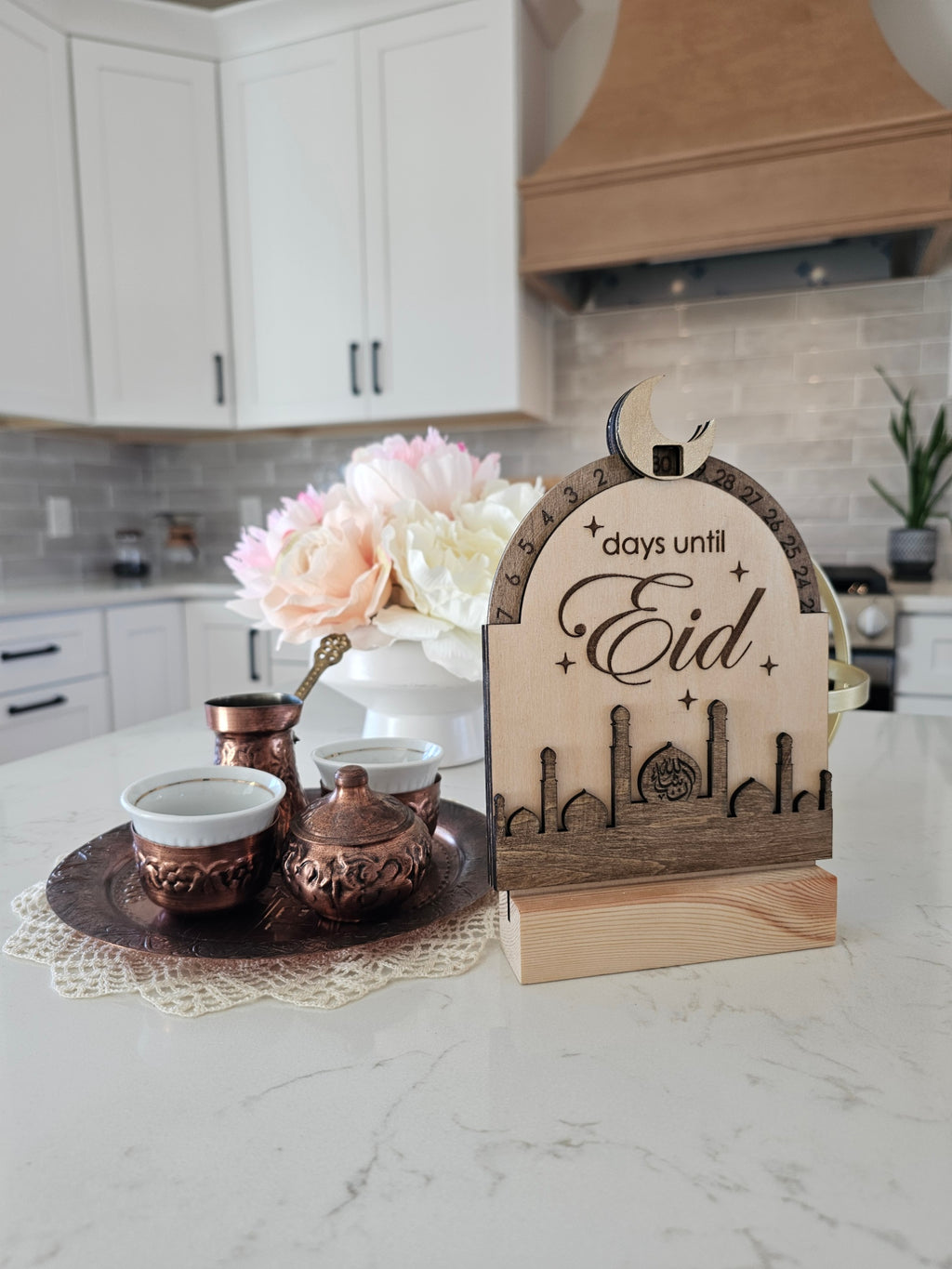 Decorative sign counting days until Eid on a kitchen counter with flowers and a teapot.