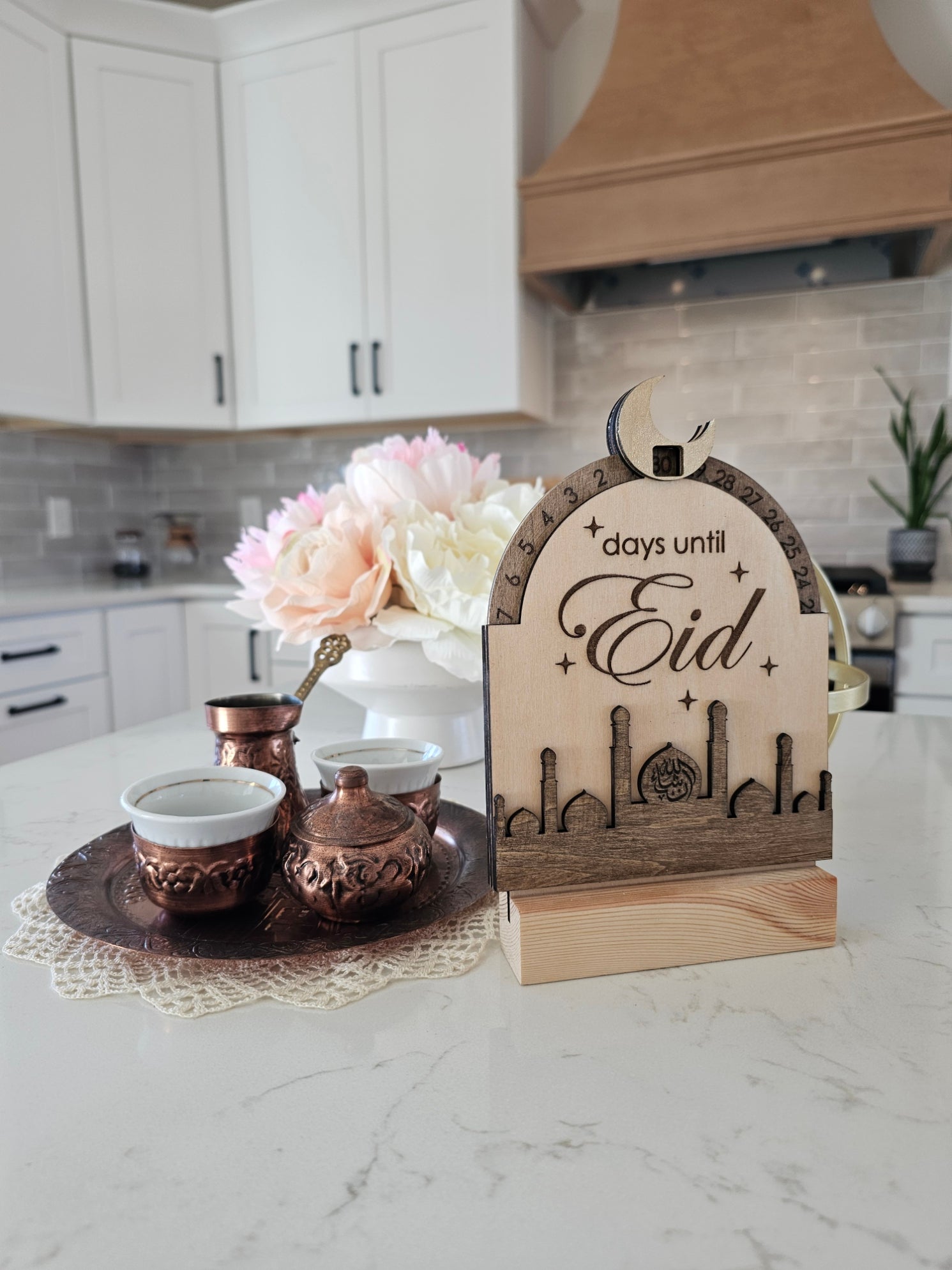 Decorative sign counting days until Eid on a kitchen counter with flowers and a teapot.